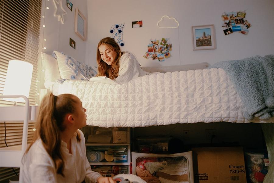 Two girls lounge on beds in dorm room.