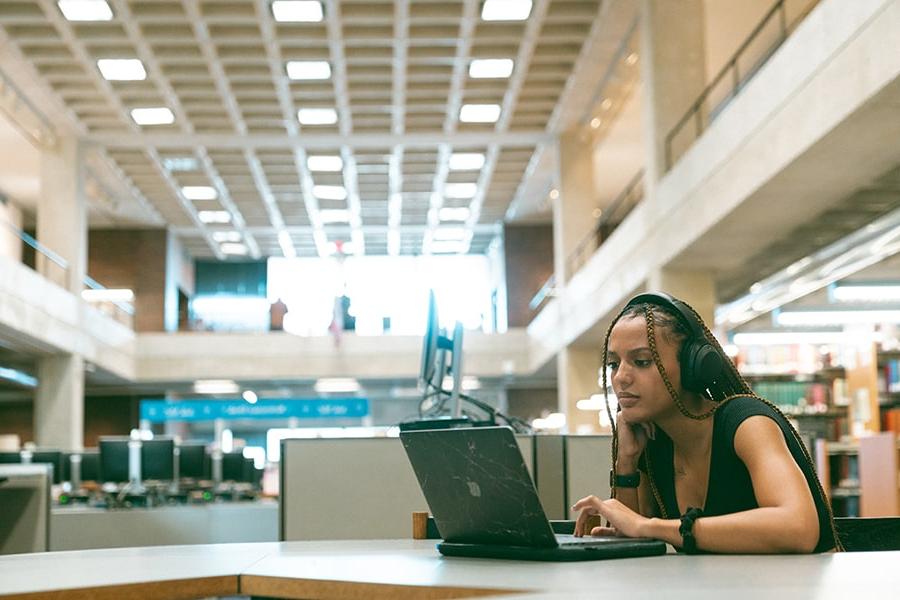 student wearing braids in library with headphones and laptop